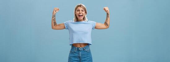 stark kvinnor stå upp och bekämpa för rättigheter. porträtt av Lycklig underhöll och Häftigt ung kvinna atletisk blond med tatueringar blinka och leende som visar muskler och biceps över blå vägg foto
