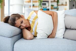 kvinna liggande på soffa ser sjuk i de levande rum. skön ung kvinna liggande på säng och innehav händer på henne mage. kvinna har smärtsam magont på säng, menstruations- period foto