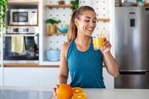 skön ung kvinna dricka färsk orange juice i kök. friska diet. Lycklig ung kvinna med glas av juice och orange på tabell i kök. foto