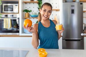 skön ung kvinna dricka färsk orange juice i kök. friska diet. Lycklig ung kvinna med glas av juice och orange på tabell i kök. foto