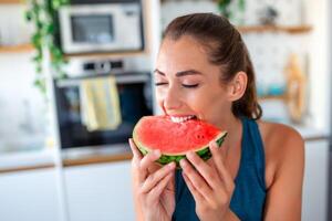 en ung kvinna är sett i henne kök, lyckligt njuter en skiva av saftig vattenmelon. henne uttryck speglar ren njutning som hon tar varje bita, förkroppsligande de enkel nöje av de ögonblick. foto