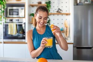 skön ung kvinna dricka färsk orange juice i kök. friska diet. Lycklig ung kvinna med glas av juice och orange på tabell i kök. foto