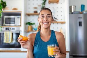 skön ung kvinna dricka färsk orange juice i kök. friska diet. Lycklig ung kvinna med glas av juice och orange på tabell i kök. foto