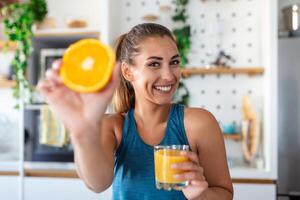 skön ung kvinna dricka färsk orange juice i kök. friska diet. Lycklig ung kvinna med glas av juice och orange på tabell i kök. foto