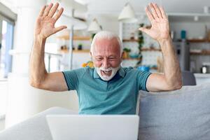 äldre man sittande på soffa ser på bärbar dator skärm skrika med glädje känner upphetsad Lycklig fira lotteri seger, tur- ögonblick, fick uppkopplad möjlighet, försäljning och rabatter e-handel begrepp foto