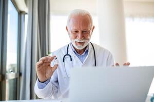 läkare uppkopplad. senior läkare talande till klient på bärbar dator dator från hans kontor. mogna manlig terapeut förklara medicinsk behandling till patient genom en video ring upp med bärbar dator i de samråd. foto