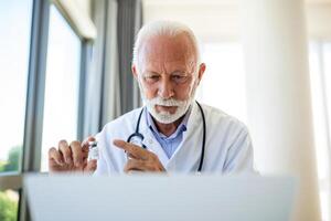 läkare uppkopplad. senior läkare talande till klient på bärbar dator dator från hans kontor. mogna manlig terapeut förklara medicinsk behandling till patient genom en video ring upp med bärbar dator i de samråd. foto