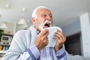senior sjuk skäggig man nyser in i servett på Hem på grå soffa med vit filt. sjukdom, skydd, coronavirus, virus, sjukdom, influensa, respiratorisk klä på sig. foto