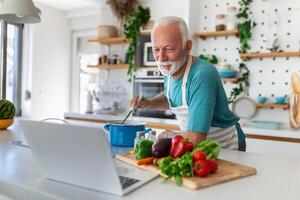 Lycklig senior man har roligt matlagning på Hem - äldre person framställning friska lunch i modern kök ser på de mottagande på hans bärbar dator foto