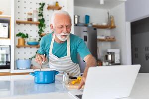 Lycklig senior man har roligt matlagning på Hem - äldre person framställning friska lunch i modern kök ser på de mottagande på hans bärbar dator foto