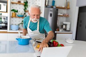 Lycklig senior man har roligt matlagning på Hem - äldre person framställning friska lunch i modern kök ser på de mottagande på hans bärbar dator foto