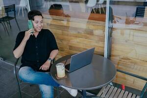 leende ung manlig affärsman talande på de telefon, Sammanträde avslappnad på Kafé tabell med bärbar dator och glas av iced mjölk kaffe. Lycklig man chattar med vänner förvirrad från arbete. foto