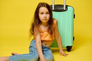 äkta caucasian liten barn flicka resande med lång hår, Sammanträde nära henne eleganta resväska, över gul bakgrund foto