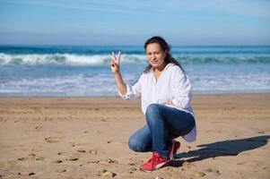 ung kvinna som visar en fred tecken ser på kamera, Sammanträde ensam på de sandig strand foto