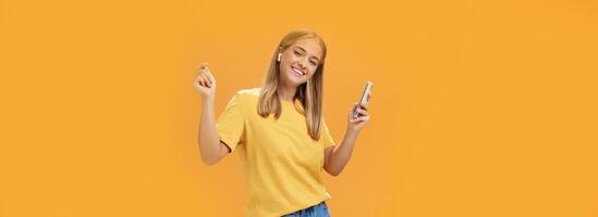 glad sorglös europeisk kvinna bär trådlös öronsnäckor innehav smartphone dans med Uppfostrad händer och nöjd Lycklig leende ledande vanlife har tid till koppla av och njut av liv över orange vägg foto