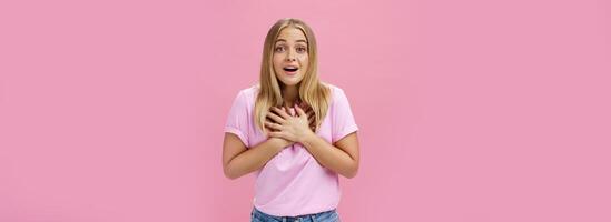 inomhus- skott av rörd överraskad snygg trevlig flicka i t-shirt och jeans innehav palmer på hjärta gasning från nöje och häpnad reagerar till grymt bra trevlig Nyheter tacksam och Lycklig foto