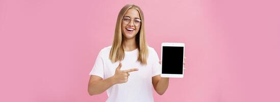 kvinna frilans programmerare stolt som visar henne app för digital läsplatta innehav grej pekande på enhet skärm leende brett med förtjust uttryck bär glasögon, Framställ över rosa vägg foto