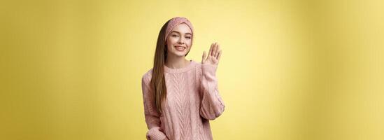 Hej äventyr. charmig glamour 20s flicka i Tröja, stickat pannband vinka handflatan ordspråk Hej vänlig leende, innehav hand i ficka gretting vänner, Framställ positiv och sorglös över gul vägg foto