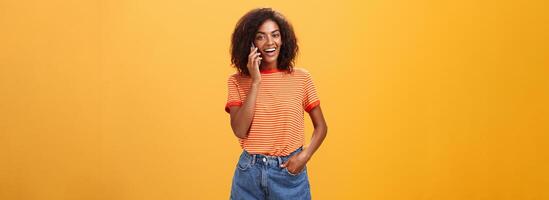 inomhus- skott av eleganta självsäker kvinna medarbetare i trendig t-shirt och denim shorts innehav smartphone nära öra kallelse vän talande via mobiltelefon vardagligt handla om flickor grejer över orange bakgrund foto