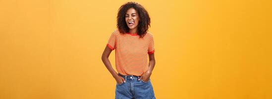 kvinna ibland vill ha känna barnslig. glad entusiastisk och karismatisk ung afrikansk amerikan kvinna med afro frisyr blinka lyckligt som visar tunga innehav händer i fickor mot orange vägg foto