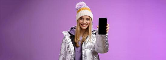 vänlig glad självsäker blond flicka i silver- eleganta vinter- jacka hatt förlänga ärm som visar smartphone visa reklam grymt bra ny enhet app leende självförsäkrad rekommendera använda sig av mobil telefon foto