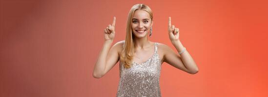 upphetsad underbar blond europeisk kvinna i silver- skinande elegant klänning höja händer pekande upp som visar imponerande otrolig annons leende lyckligt glada vill ta närmare se, röd bakgrund foto
