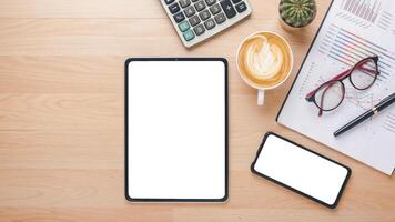 topp se av en trä- skrivbord terar en tom läsplatta skärm, smartphone, kalkylator, och en latte konst kaffe, vid sidan av företag diagram och glasögon. foto