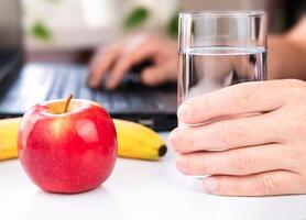 en mannens hand innehar en glas av vatten och ett äpple och en banan närliggande. friska mellanmål på arbete. närbild. selektiv fokus. foto