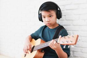 asiatisk pojke bär hörlurar med svart musik sitta och spela gitarr i de hus. musik inlärning begrepp, musik skicklighet Träning foto