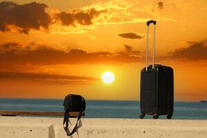 resväska och väska är stående på en strand med en solnedgång i de bakgrund.resor och Semester begrepp foto