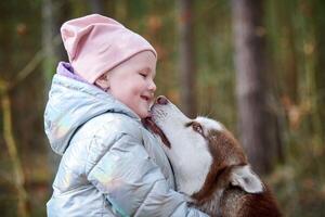 söt liten flicka i rosa hatt och ljus blå jacka kramar sibirisk hes hund och leende foto