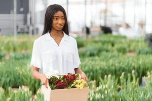 skön ung leende afrikansk amerikan flicka, arbetstagare med blommor i växthus. begrepp arbete i de växthus, blommor. foto