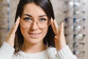 kvinna väljer glasögon i de Lagra. brunett i en vit Tröja köper glasögon. flicka på en bakgrund av affär fönster med annorlunda modeller av glasögon foto