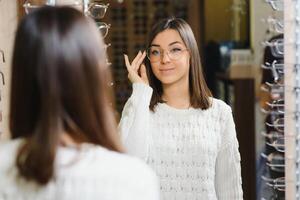 kvinna väljer glasögon i de Lagra. brunett i en vit Tröja köper glasögon. flicka på en bakgrund av affär fönster med annorlunda modeller av glasögon foto