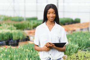 skön ung leende afrikansk amerikan flicka, arbetstagare med blommor i växthus. begrepp arbete i de växthus, blommor. foto