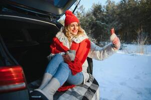 ung kvinna Sammanträde i bil trunk dricka värma te och tar en selfie på vinter- snöade dag. foto