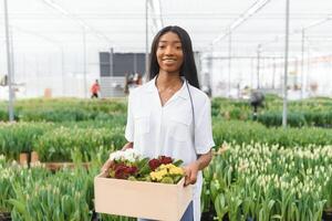 skön ung leende afrikansk amerikan flicka, arbetstagare med blommor i växthus. begrepp arbete i de växthus, blommor. foto