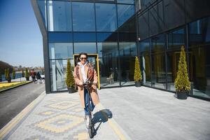 ung skön kvinna i en jacka ler och rider ett elektrisk skoter till arbete längs kontor byggnader foto