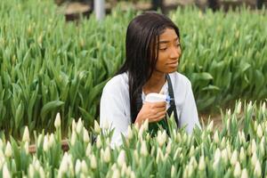 skön ung leende afrikansk amerikan flicka, arbetstagare med blommor i växthus. begrepp arbete i de växthus, blommor. foto