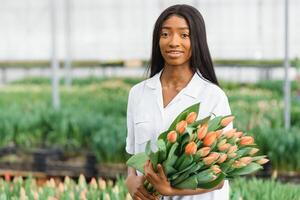 skön ung leende afrikansk amerikan flicka, arbetstagare med blommor i växthus. begrepp arbete i de växthus, blommor. foto