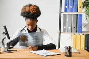 kvinna afrikansk amerikan läkare bär vit täcka med stetoskop Sammanträde Bakom skrivbord i de kontor och ser röntgen bild. foto