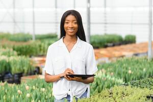 skön ung leende afrikansk amerikan flicka, arbetstagare med blommor i växthus. begrepp arbete i de växthus, blommor. foto