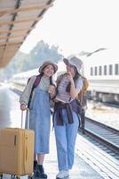 två ung asiatisk vänner flickor med ryggsäckar på järnväg station väntar för tåg, två skön kvinnor gående längs plattform på tåg station foto