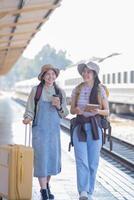 två ung asiatisk vänner flickor med ryggsäckar på järnväg station väntar för tåg, två skön kvinnor gående längs plattform på tåg station foto