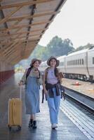 två ung asiatisk vänner flickor med ryggsäckar på järnväg station väntar för tåg, två skön kvinnor gående längs plattform på tåg station foto