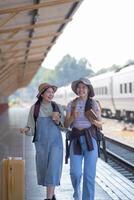 två ung asiatisk vänner flickor med ryggsäckar på järnväg station väntar för tåg, två skön kvinnor gående längs plattform på tåg station foto