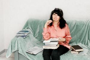 trött senior kvinna på de soffa omgiven förbi böcker och anteckningar foto
