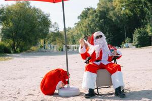 santa claus i solglasögon drycker en cocktail medan liggande på en Sol vilstol på de sjö strand. santa i de tropikerna foto