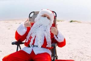 santa claus tar bort en medicinsk mask liggande på en Sol vilstol på de strand. slutet av de epidemi foto