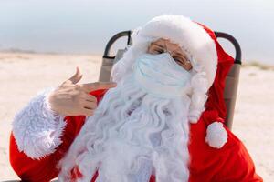 santa claus tar bort en medicinsk mask liggande på en Sol vilstol på de strand. slutet av de epidemi foto